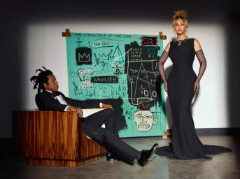 Beyonce standing in a long black dress and Jay-Z seated in a wooden armchair before a teal abstract painting