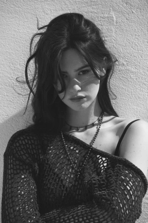 Portrait of a woman with dark hair wearing a crocheted top and necklaces against a textured wall.