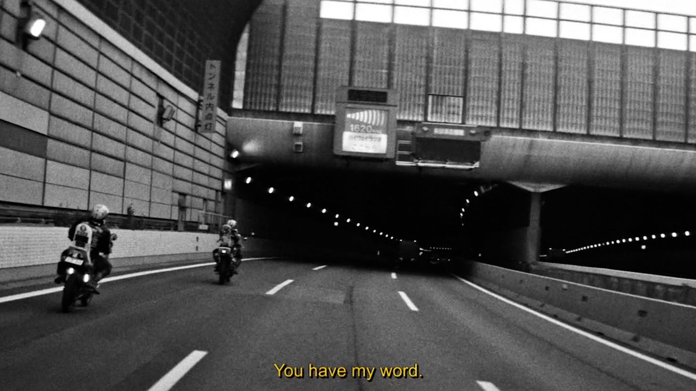 Two motorcyclists ride through an urban tunnel on a city road in a monochrome image, captured at eye-level with visible overhead lighting.