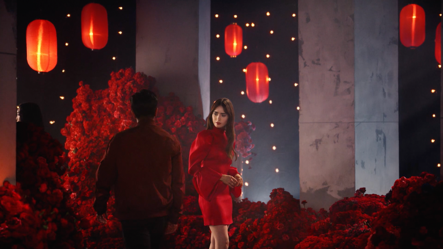 A stylish woman in a red dress poses in a studio with flowers, lanterns, and a man, set against a backdrop of moody lighting and concrete textures.