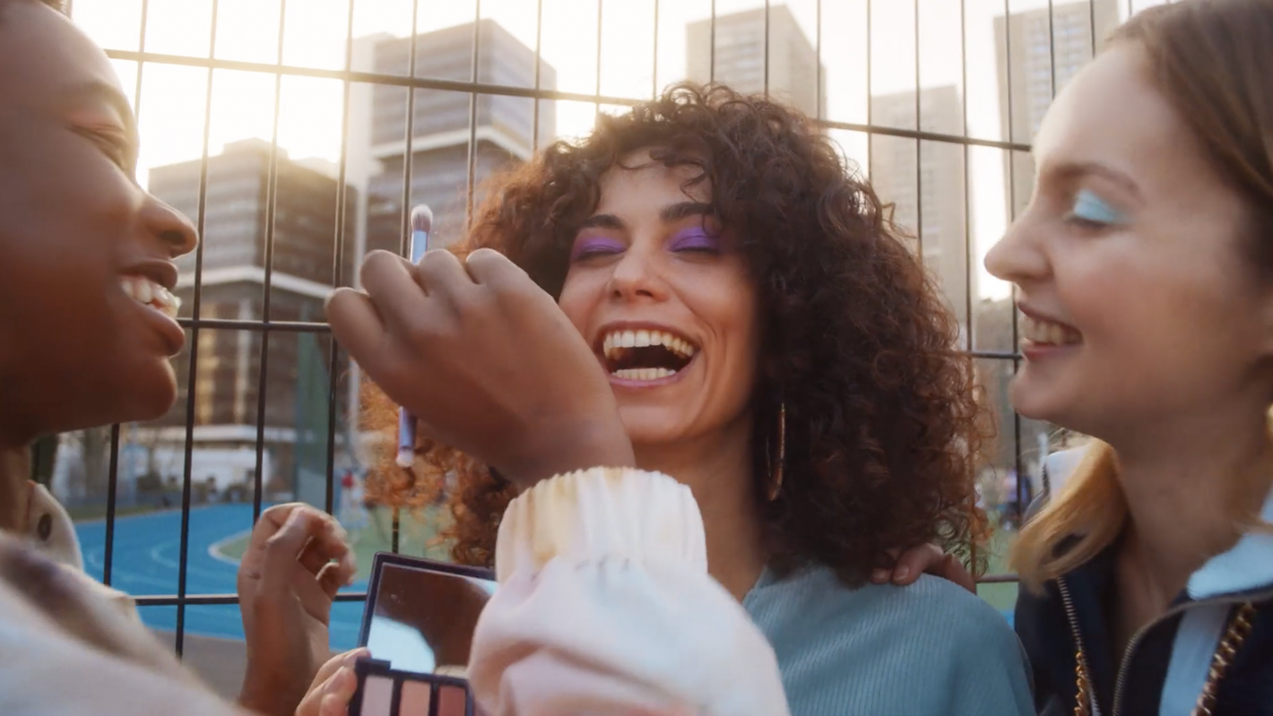 Three happy women apply colorful eyeshadow in an urban setting with tall buildings, sharing beauty and friendship in outdoor daylight.