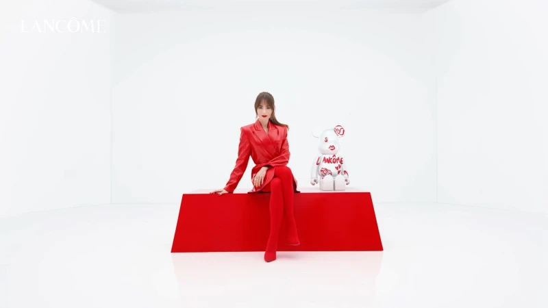 Adult woman in a red suit sits on a red platform next to a white bear statuette decorated with Lancôme logos in a bright photo studio.