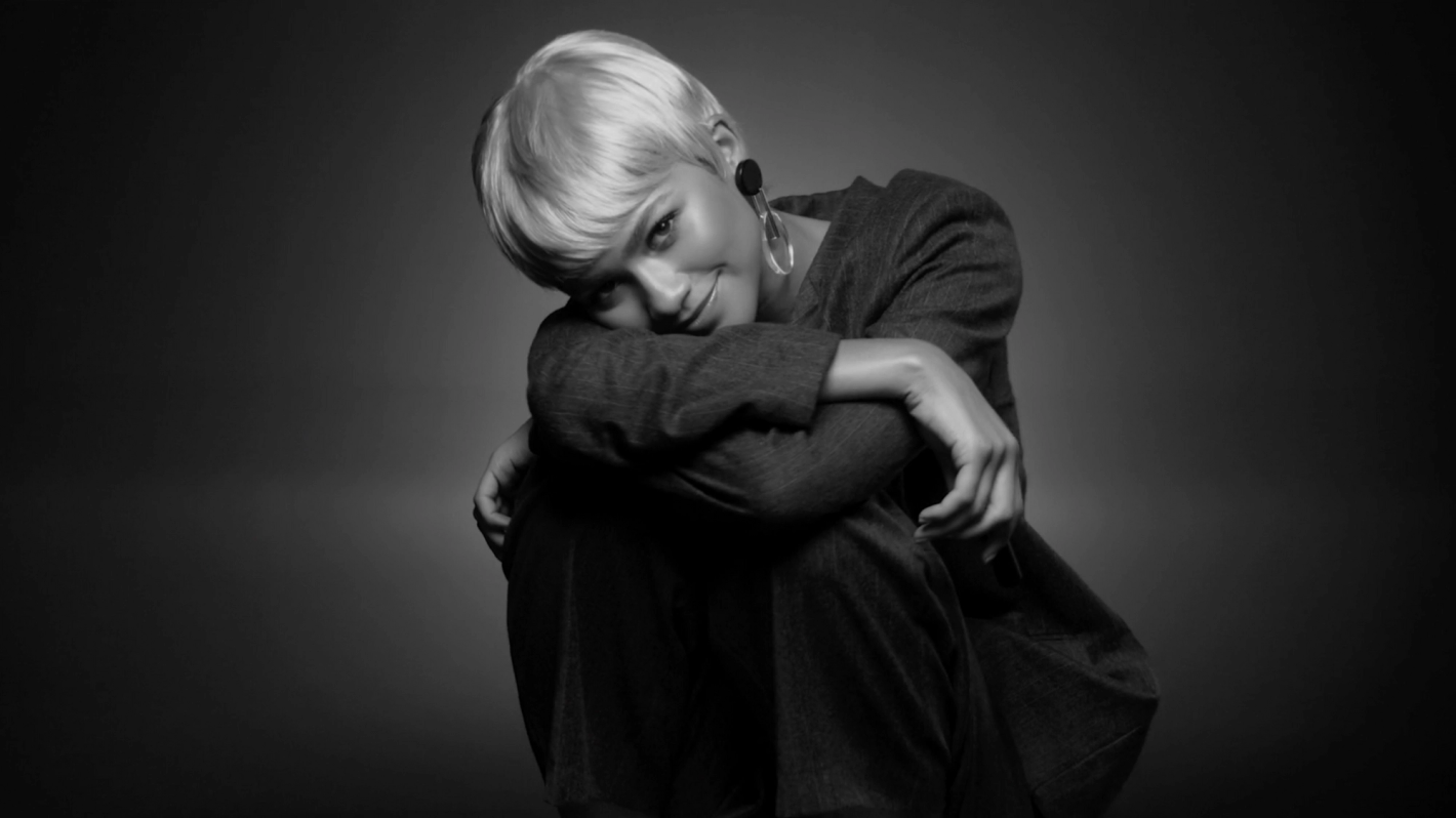 Black and white portrait of a woman with short blonde hair in a dark jacket, posing in an indoor studio against a graduated background.