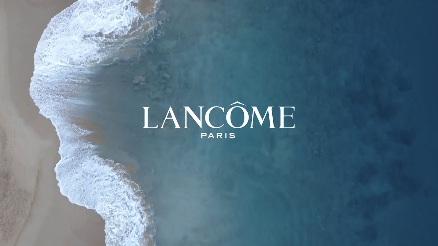 Aerial view of ocean waves crashing on sandy beach with Lancôme Paris logo in the middle, creating a serene and luxurious visual.