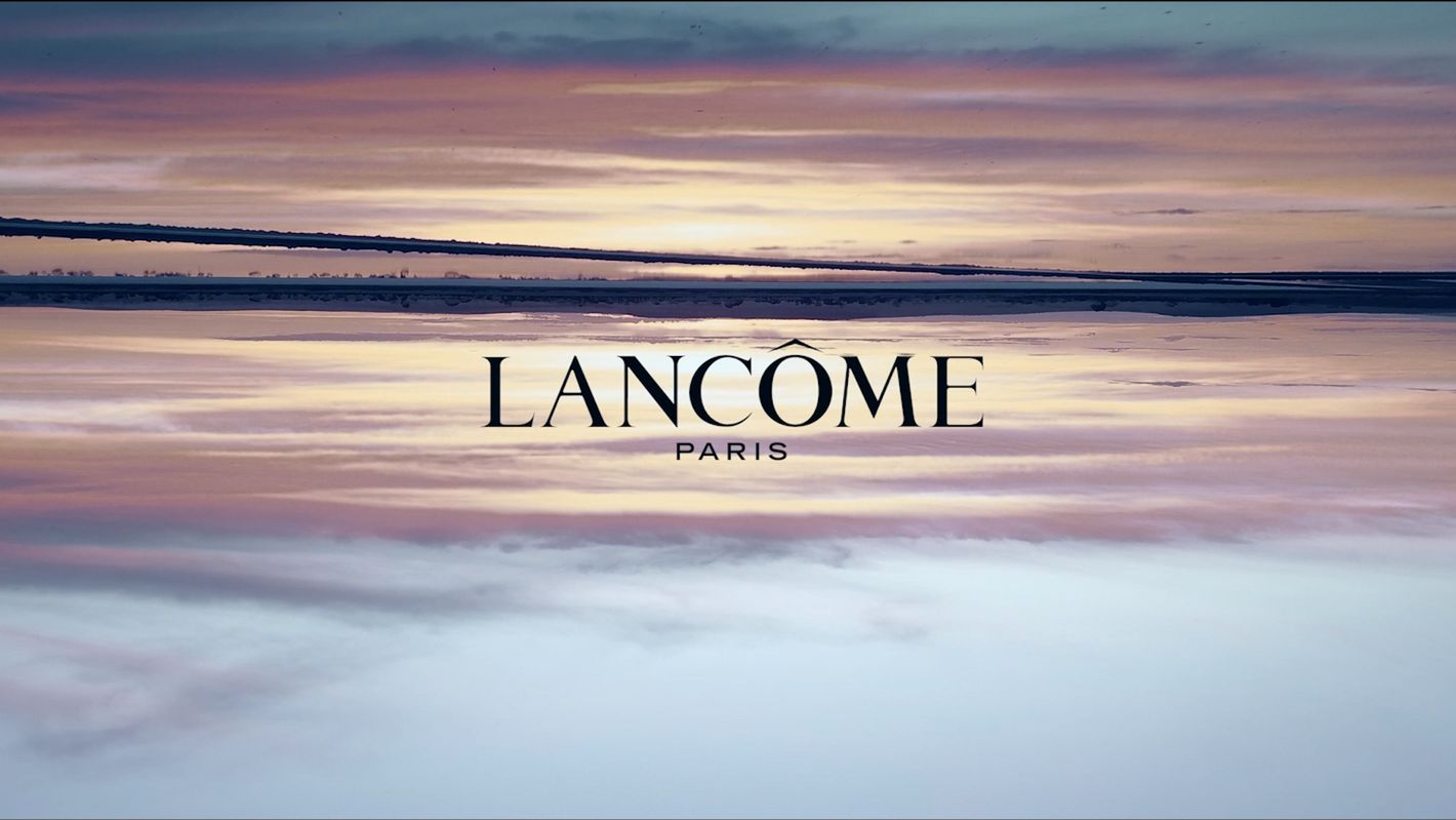 Lancôme Paris brand logo against a soft purple and orange sky reflected in the water, creating a serene and upscale beauty image.
