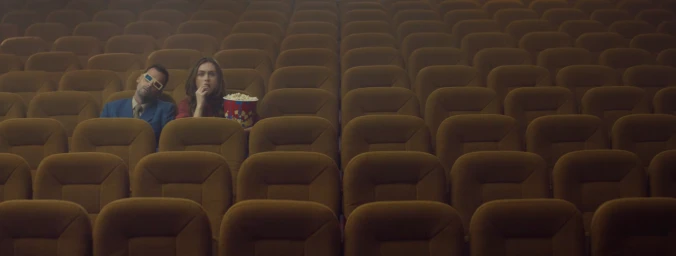 Man and woman seated in a nearly empty cinema hall with rows of brown plush seats, holding a bucket of popcorn.