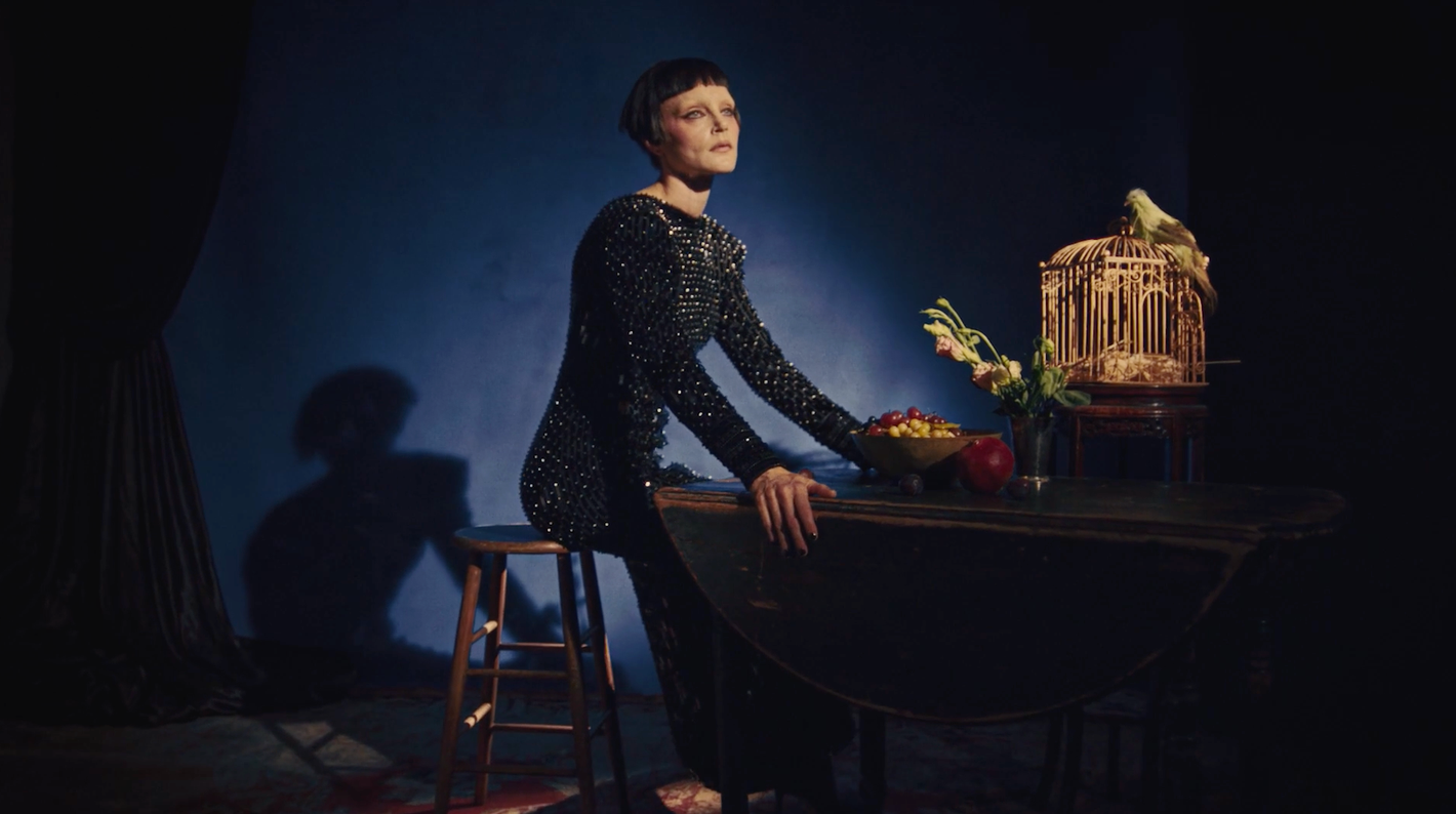 Elegant woman with short dark hair posing by a table with a birdcage, fruit, and flowers in a dimly lit room with a blue backdrop.