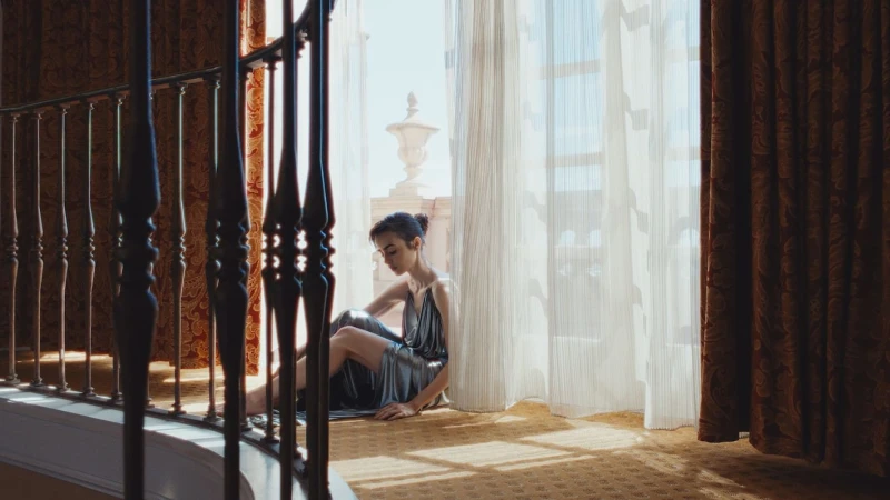 Woman in shiny silver dress seated on a hotel floor near sheer curtains by Max Bartick