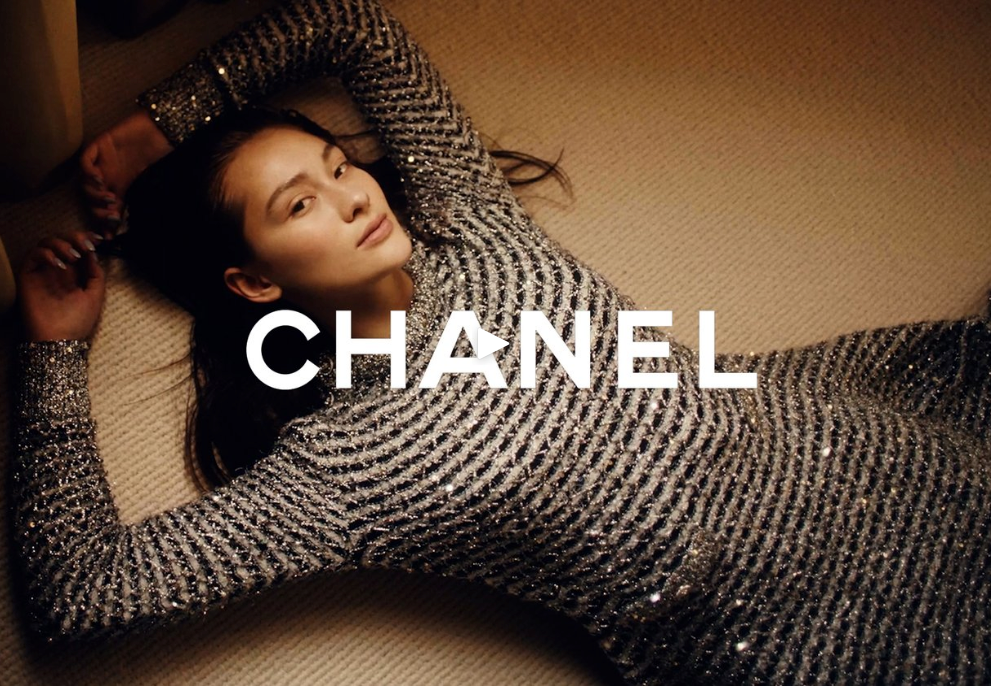 Asian woman in Chanel dress lays on carpet looking at camera. Fashion editorial shot with soft lighting, neutral tones, and visible logo.