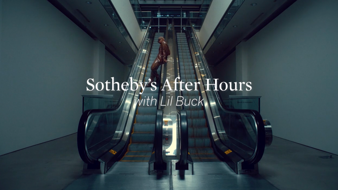 Man in a brown suit stands on an escalator in a modern building with white text overlayed on the scene, Sotheby's After Hours with Lil Buck.
