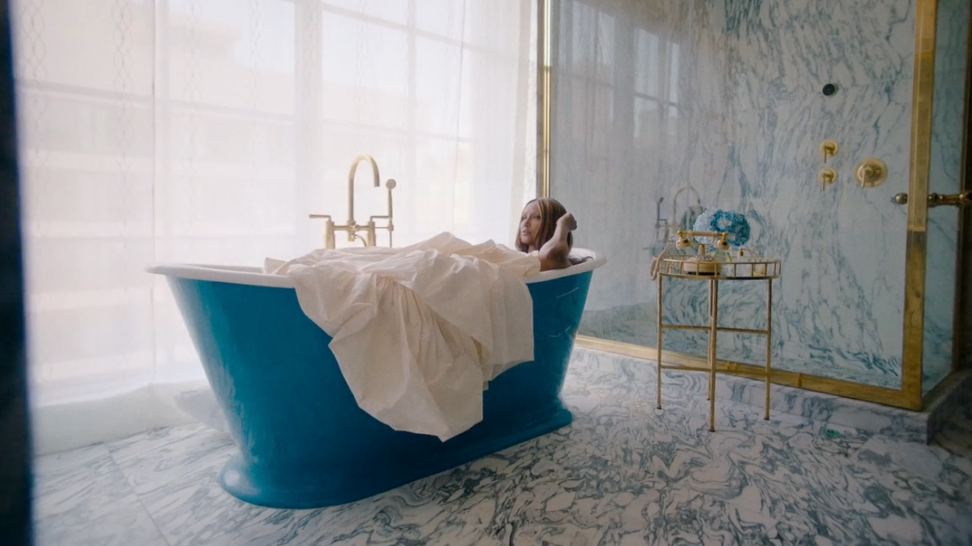 Woman relaxes in a blue bathtub, partially covered by a white cloth, within an opulent bathroom featuring marble walls and gold fixtures.