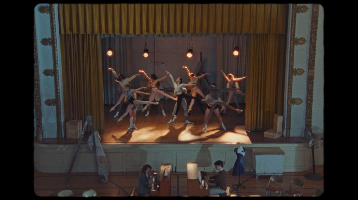 Ballet dancers perform on stage in a vintage-style theater. Additional people are sitting in front of the stage area. Golden drapes backdrop.