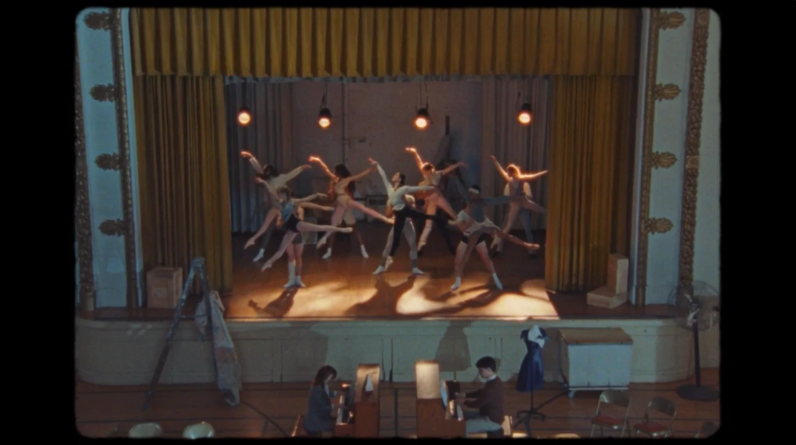 Ballet dancers perform on stage in a vintage-style theater. Additional people are sitting in front of the stage area. Golden drapes backdrop.