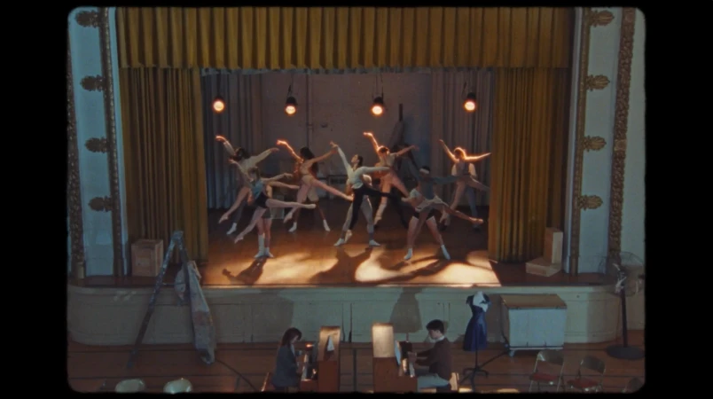 Ballet dancers perform on stage in a vintage-style theater. Additional people are sitting in front of the stage area. Golden drapes backdrop.