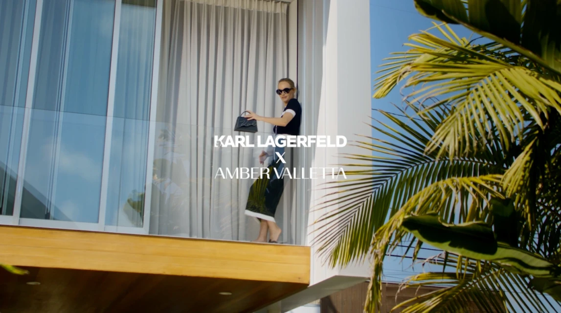 Amber Valletta stands on a balcony, wearing sunglasses, holding a black handbag, in front of palm trees and a building.
