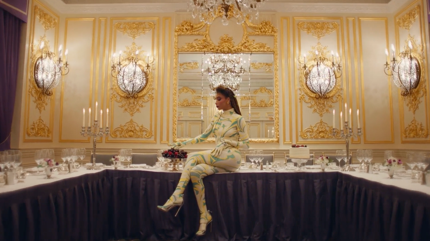 Woman in patterned catsuit sits on formal dining table in ornately decorated room with gilded details, crystal chandeliers, and set table.