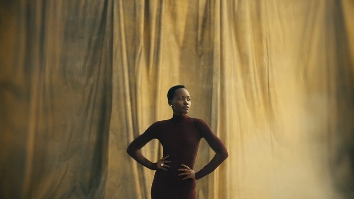 Portrait of a woman with short hair in a maroon dress standing against a textured gold backdrop. She has her hands on her hips.