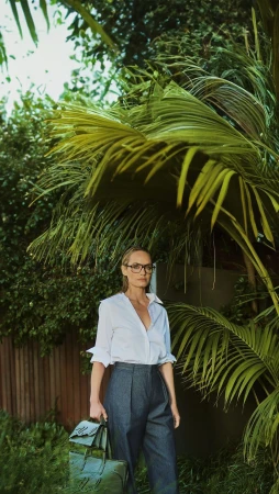 Woman in white shirt and dark trousers standing outdoors beneath palm fronds holding two green leather bags