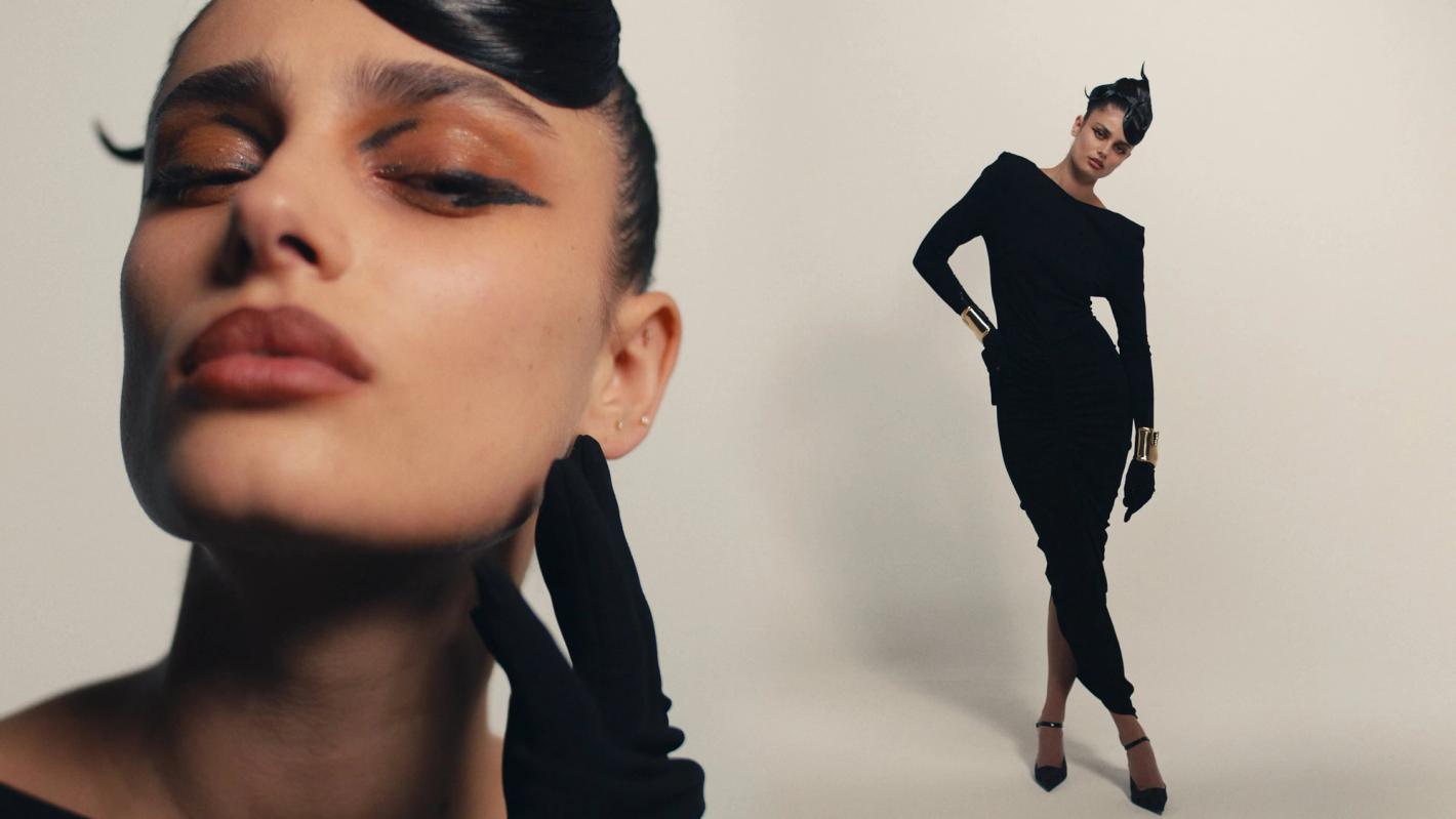 A woman with stylized makeup and hair poses in a long-sleeved black dress with gold cuffs against a plain light background.