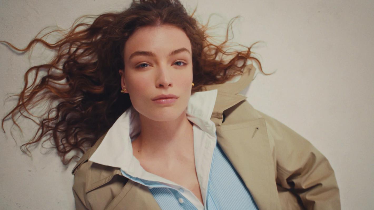 Portrait of a woman with auburn hair wearing a trench coat and collared shirt, lying on a white background in a close-up studio shot.