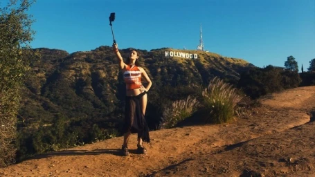 Woman taking a selfie with a phone on a stick on a dirt path with the Hollywood Sign visible in the background.