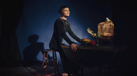 Woman in sequined black dress seated on stool at dark wooden table with fruit bowl and birdcage by Max Bartick
