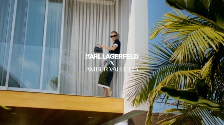 Amber Valletta stands on a glass balcony holding a handbag in a campaign directed by Max Bartick.