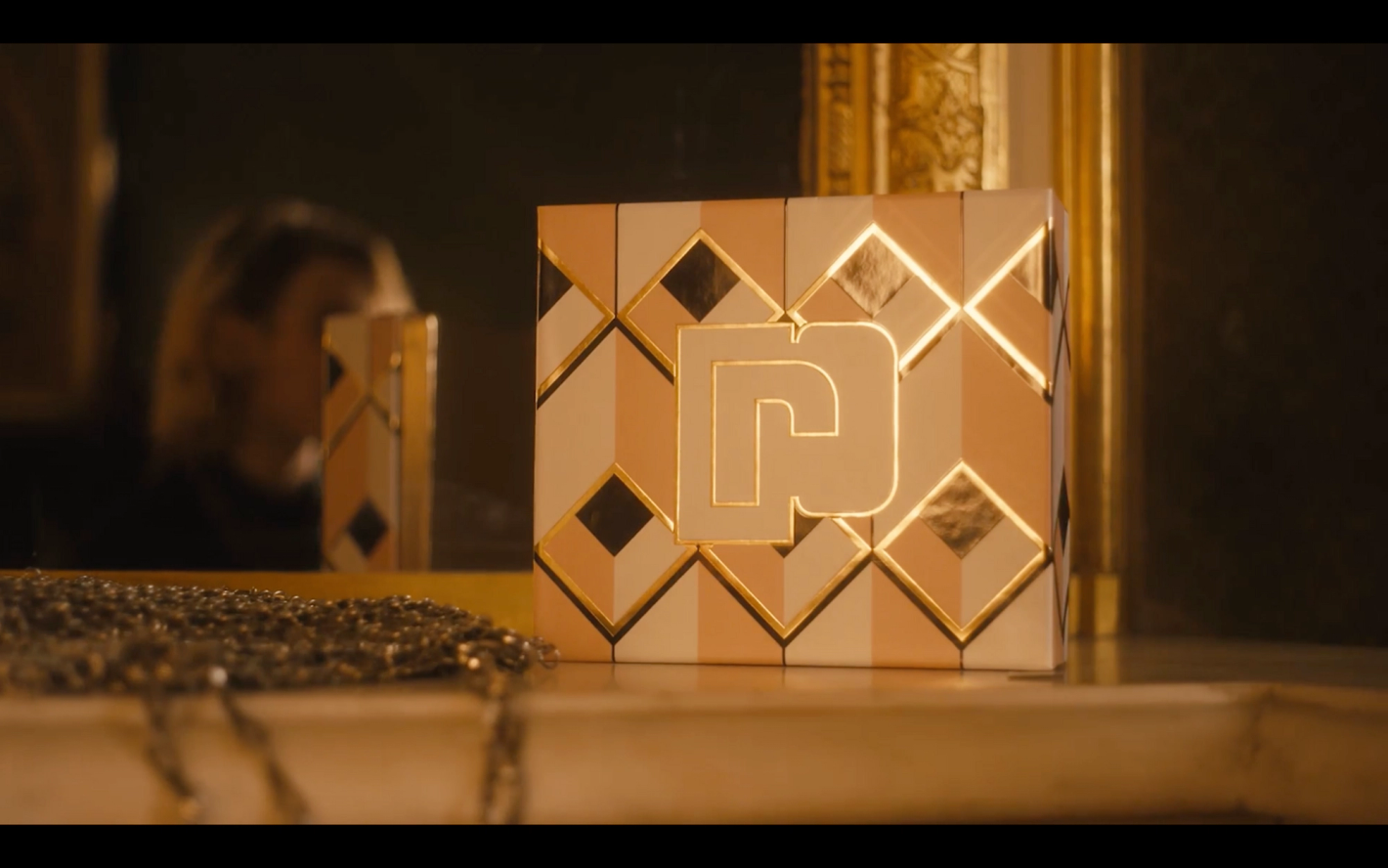Close-up of a Patou perfume box with geometric patterns and a gold logo on a light-colored surface, elegant and modern.