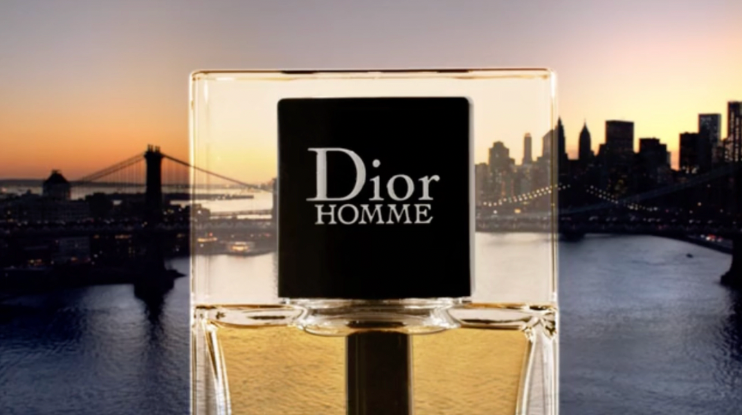 Dior Homme fragrance bottle in front of an urban skyline with a bridge over water and a setting sun background.