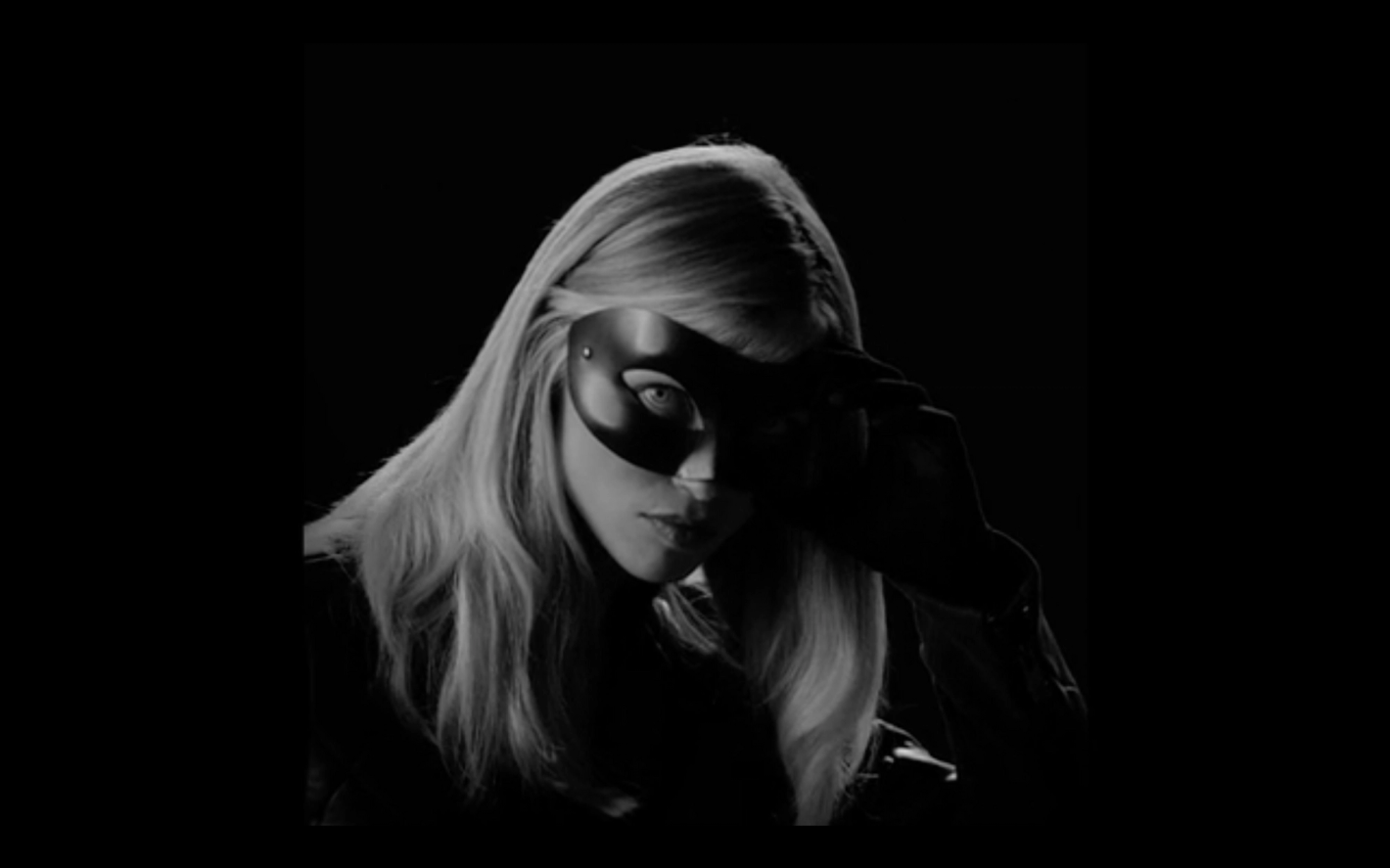 Striking monochrome portrait of a blonde woman with dark mask, face partially obscured, against a stark black background in shadow.
