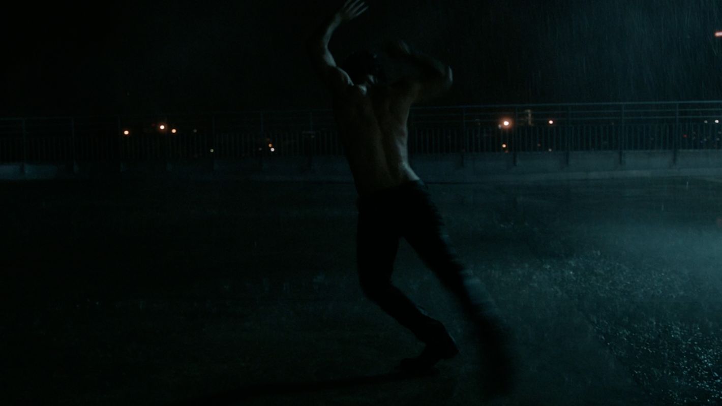 Shirtless man dancing in the rain at night in an urban environment with dark tones and soft city lights in the background.