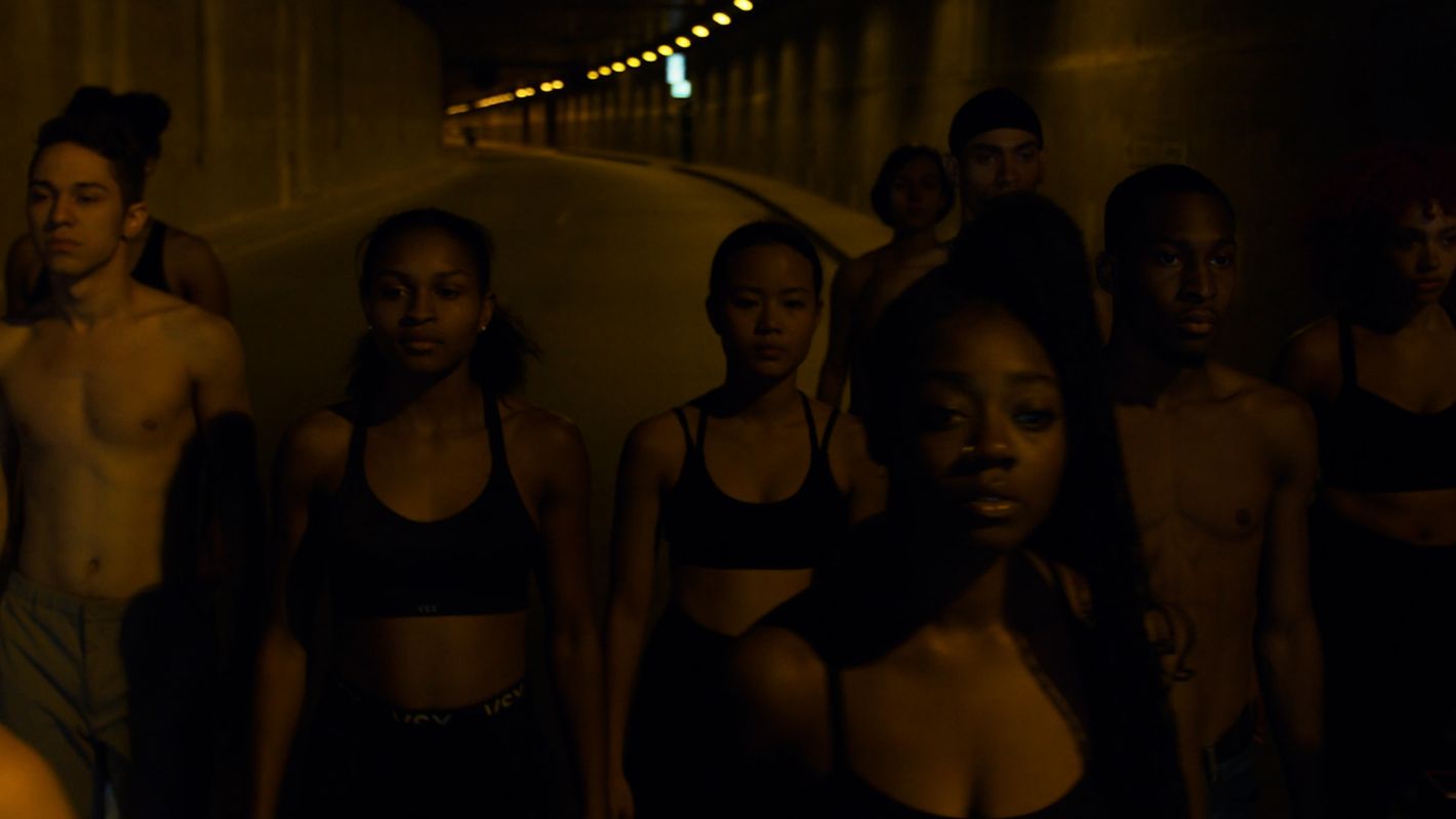 A group of diverse young adults stand in a dimly lit tunnel, shot at eye-level. Some are shirtless and wearing sportswear.