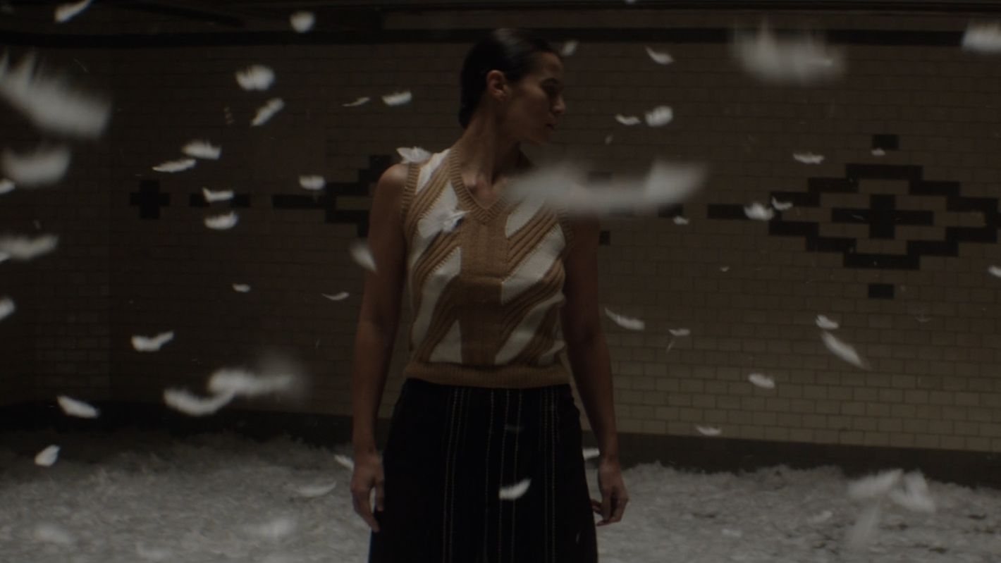 Woman in patterned top and dark skirt stands among floating feathers in an interior setting with a brick wall and dark ceiling.