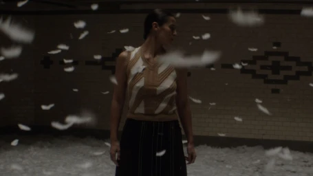 Woman in patterned knit vest standing amid falling white feathers inside a tiled underground space