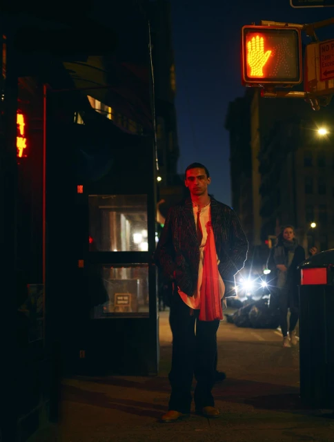 Stylish adult male stands on city street at night, wearing dark blazer, white collared shirt, dark pants, and tan footwear.