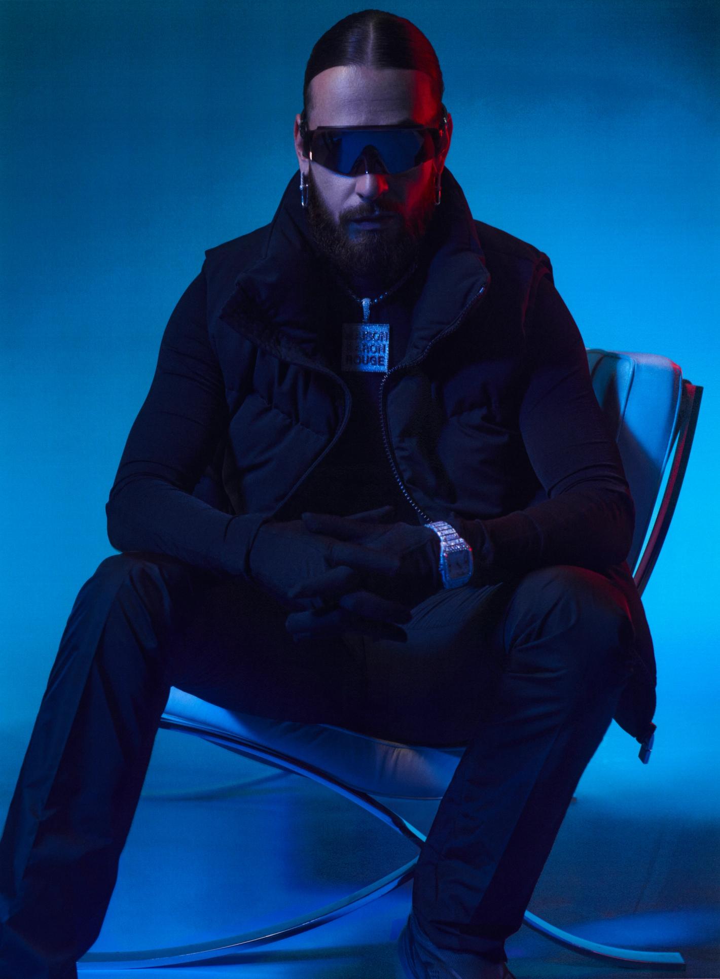 Man with beard and sunglasses in a black outfit sitting in a chair in a blue illuminated photo studio. Posing in a seated position.