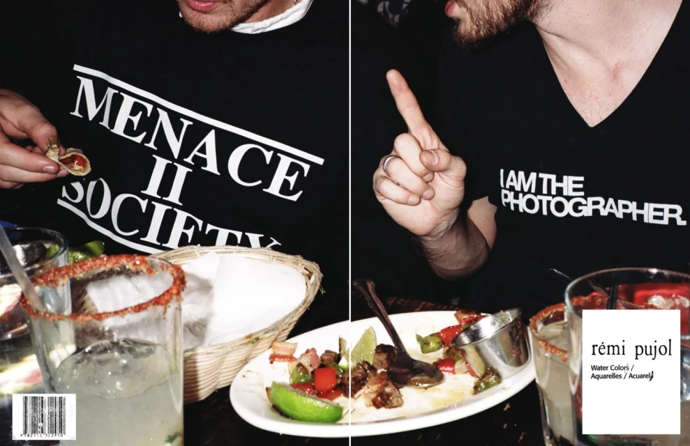Two adult males are at a restaurant with cocktails and plates of food; one is wearing a Menace II Society tee, the other a photographer tee.