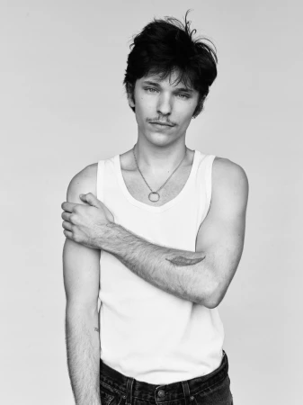 Portrait of a man in a white tank top and dark jeans posing against a plain background