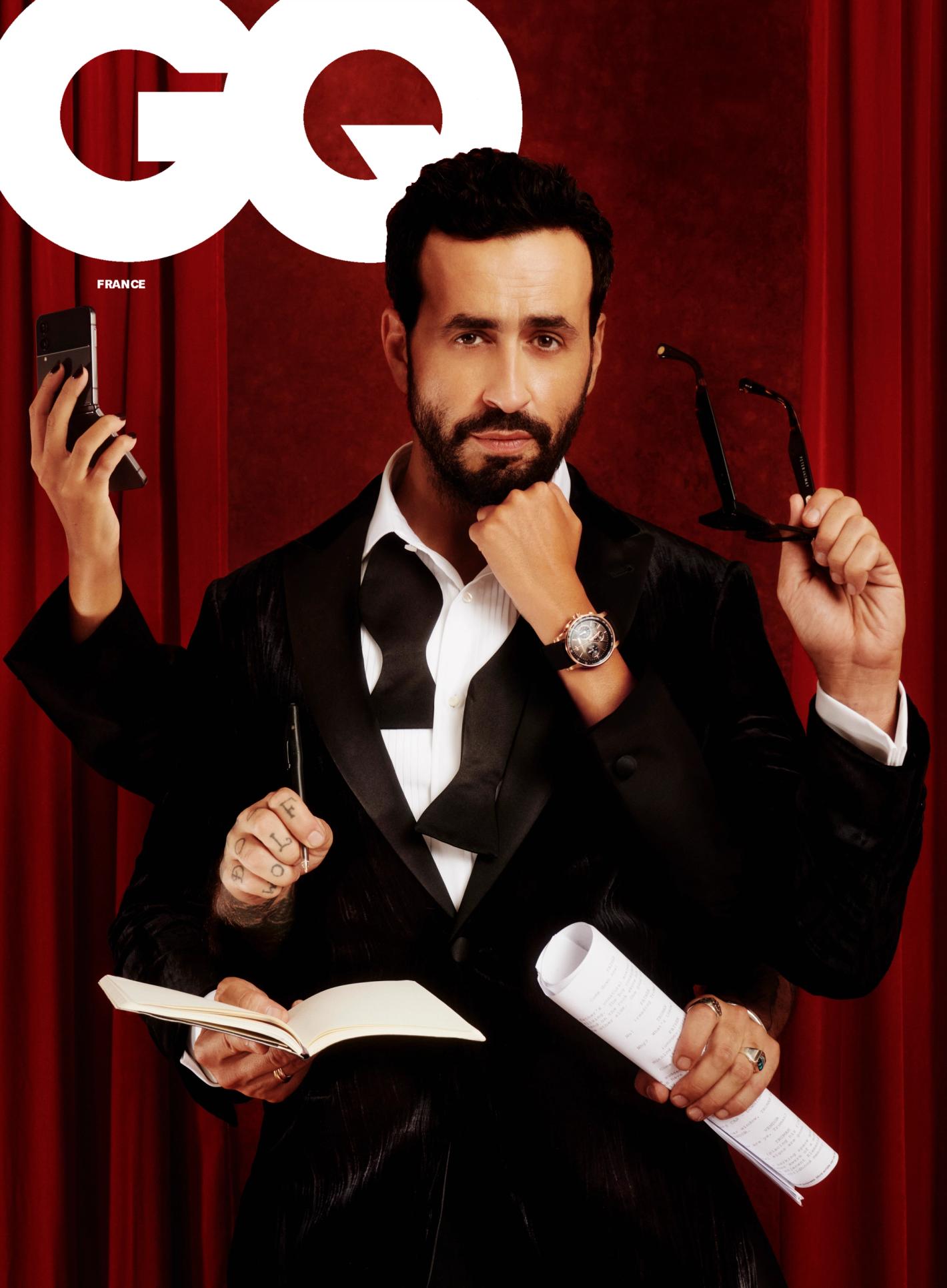 Man on GQ France cover with multiple arms holding a phone, glasses, book, and document, wearing a tuxedo against a red curtain background.