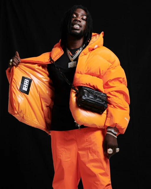 Full body studio shot of an adult Black man in an orange puffer jacket, black t-shirt, and matching pants, accessorized with jewelry and a black crossbody bag.