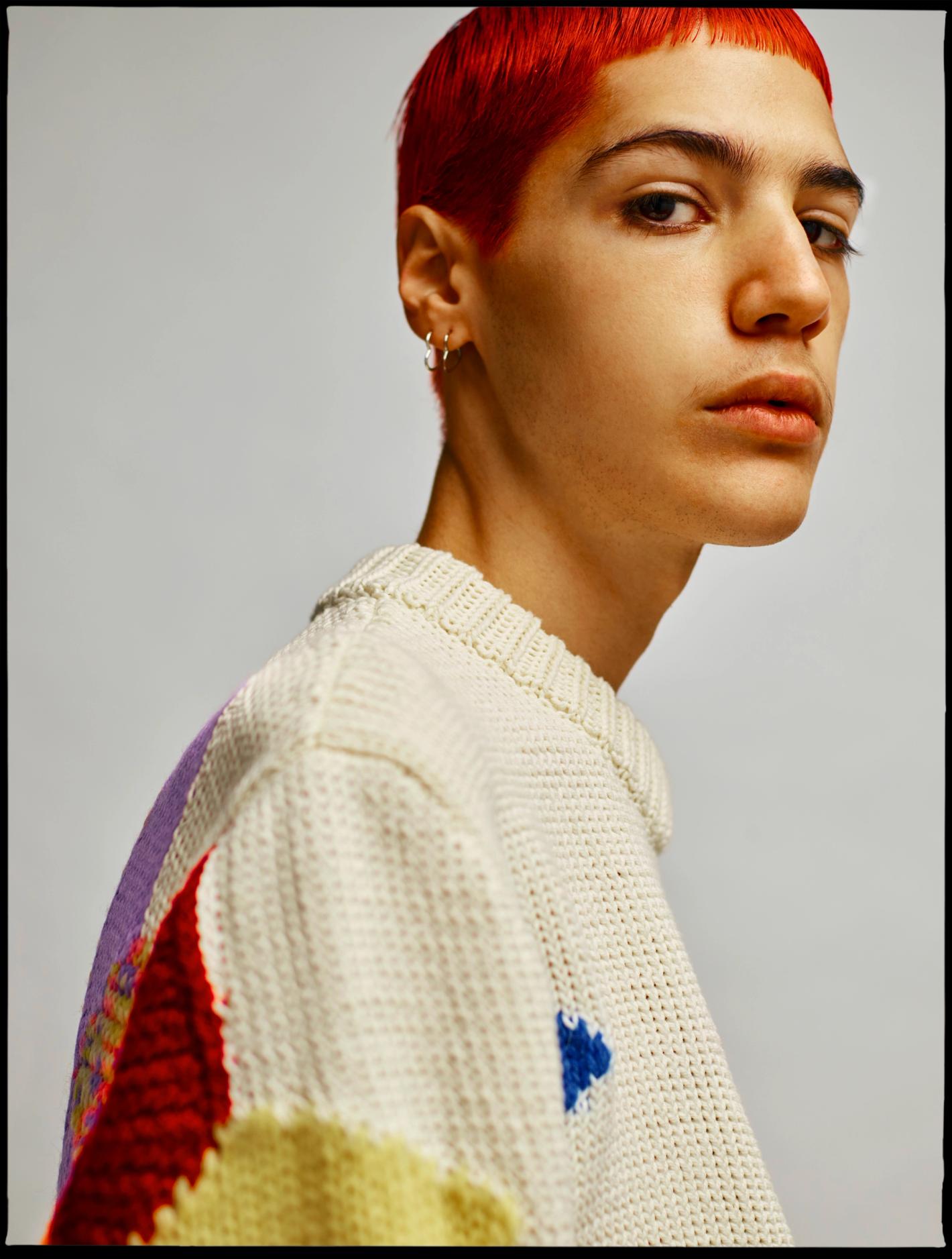 Close-up studio portrait of a young adult with dyed red short hair wearing a textured white sweater against a neutral grey backdrop.