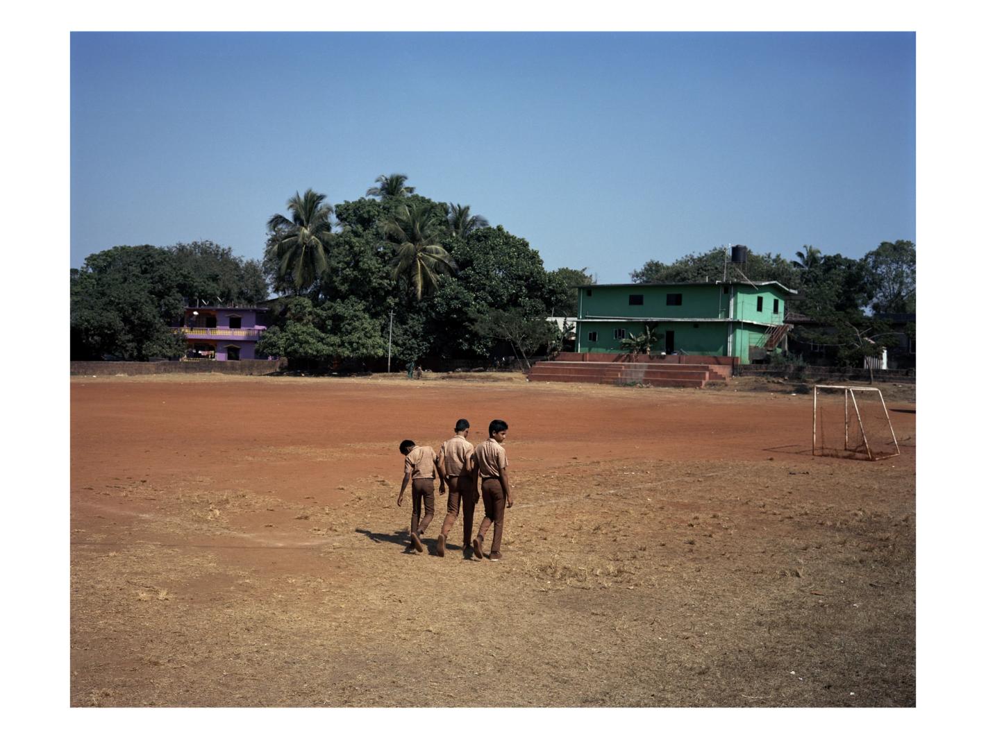 Three children in school uniforms walk across a large, dusty field in front of green trees and buildings under a clear blue sky.