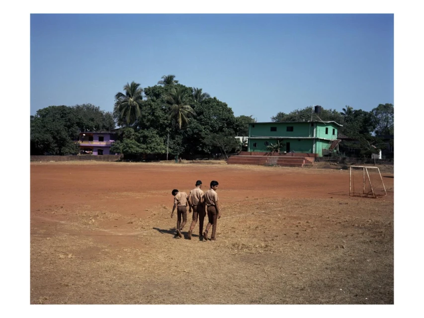 Three children in school uniforms walk across a large, dusty field in front of green trees and buildings under a clear blue sky.