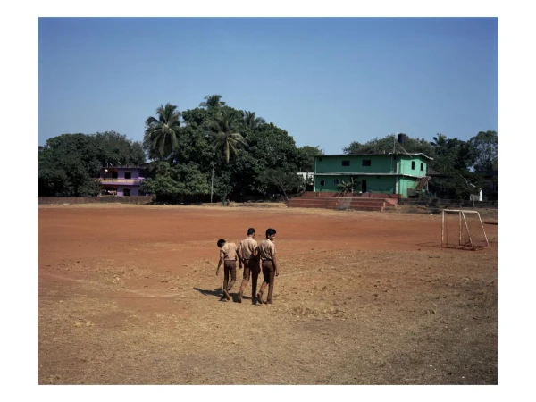 Three children in school uniforms walk across a large, dusty field in front of green trees and buildings under a clear blue sky.