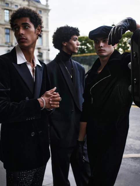 Three stylish men in dark suits pose outdoors in a European city, showcasing formal fashion and classic tailoring with a vintage aesthetic.