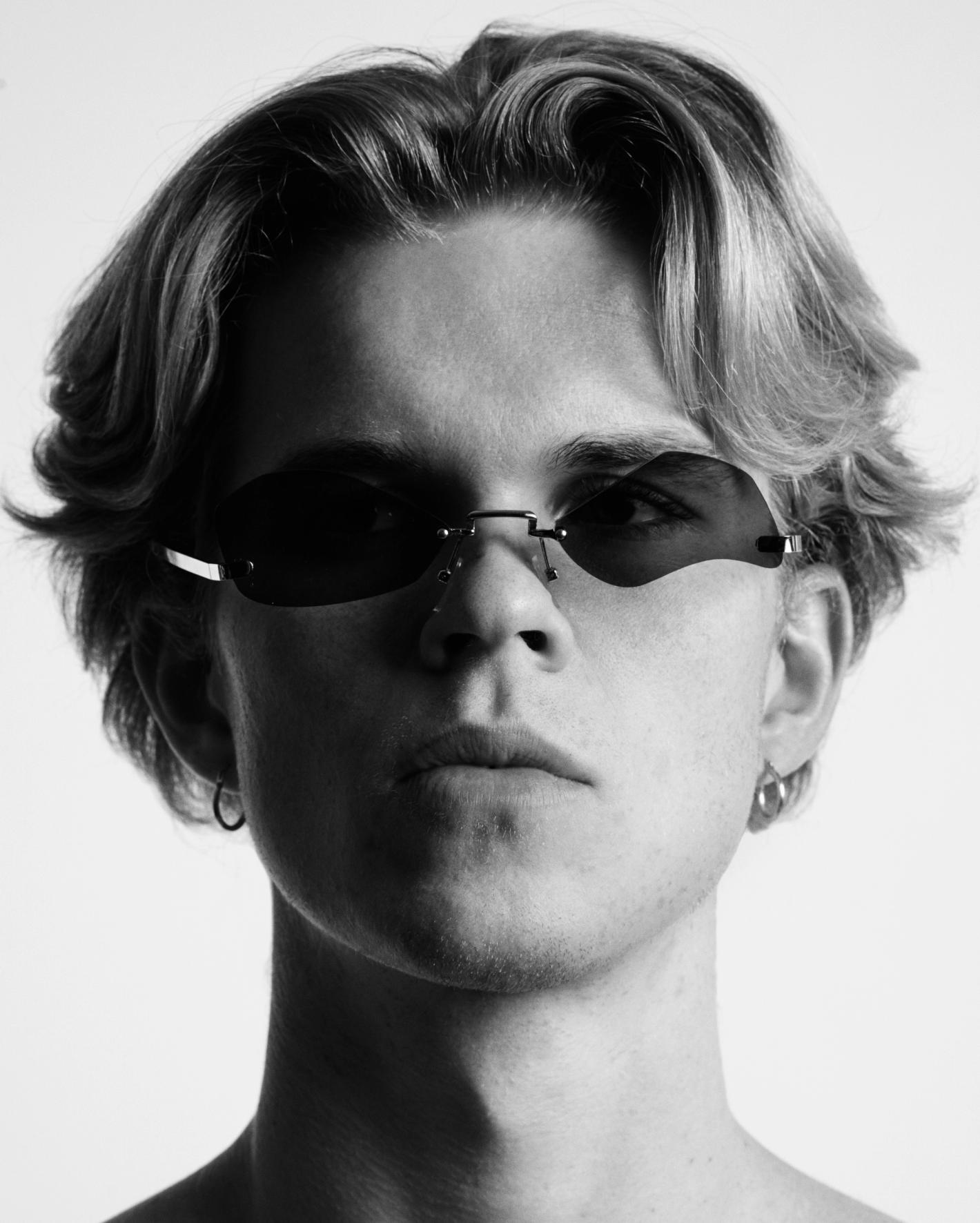 Black and white portrait of a young adult male with blonde hair wearing dark sunglasses and hoop earrings against a white backdrop.