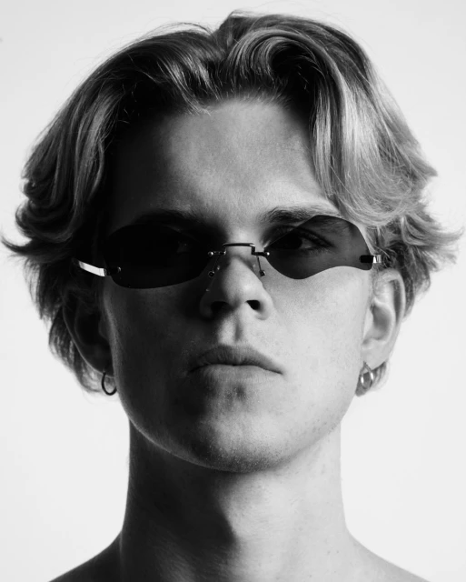 Black and white portrait of a young adult male with blonde hair wearing dark sunglasses and hoop earrings against a white backdrop.