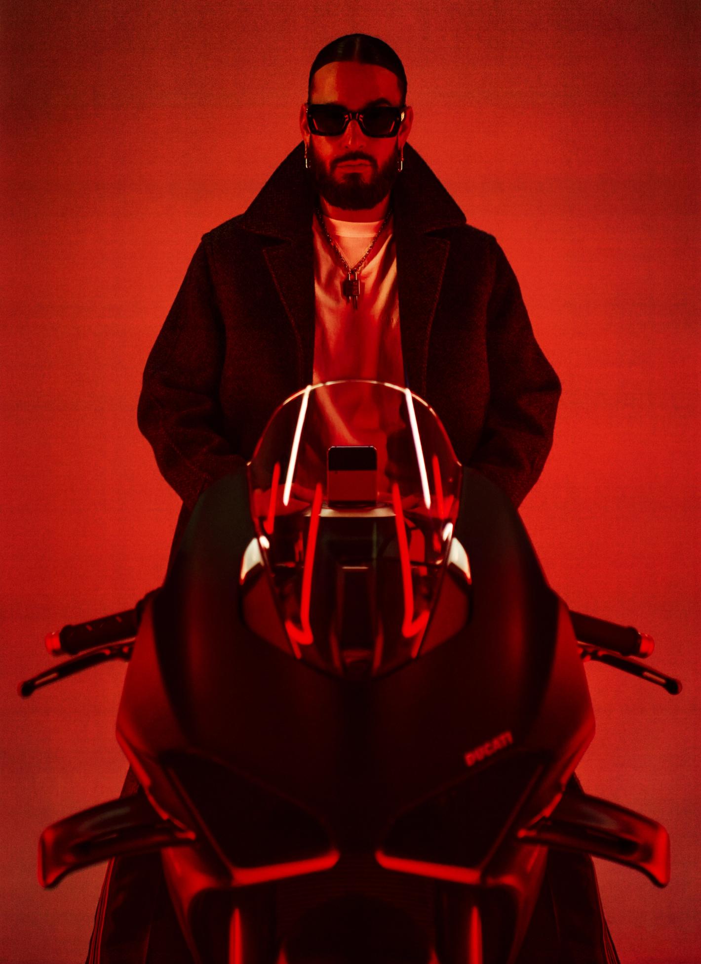 Portrait of a man with a beard and sunglasses, wearing a black coat, sitting on a motorcycle in a red photo studio setting.