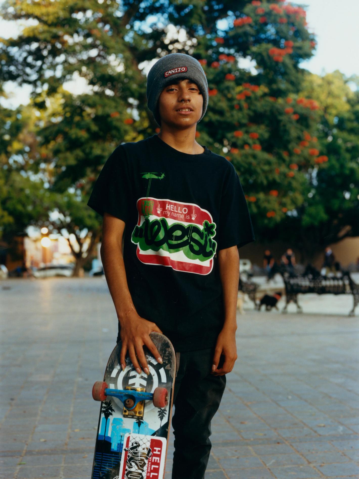 Teen skater poses outdoors in a black t-shirt and beanie, holding a skateboard against a backdrop of a public square and leafy trees.