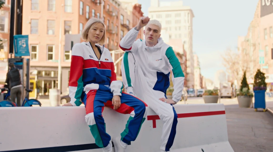 Two people in tracksuits sit on a concrete barrier in an urban setting with buildings and trees in the background.